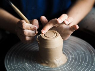 Ceramist at work using tools for creating handmade vase in studio, selective focus. Close up of female potter hands sharing handcraft crockery in studio. Small business, art, hobby concept.