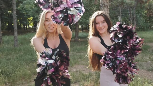 Two beautiful cheerleader woman dancing with pompons outdoors in the park on sunset