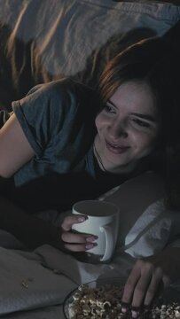 Comedy show. Insomnia night. Home leisure. Happy woman watching funny movie eating pop corn drinking beverage laying bed dark room interior. Vertical video.