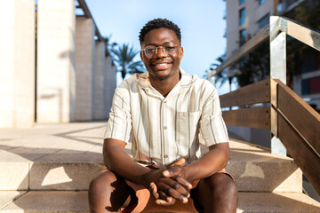Happy African American young man sitting on stairs outdoors looking at camera.