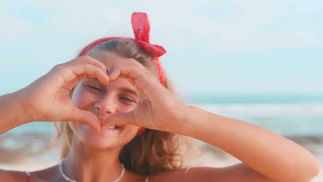 Young friendly beautiful Caucasian woman teenager shows heart from fingers to camera sending video valentine to boyfriend or close friends poses with smile stands on sunny beach near sea