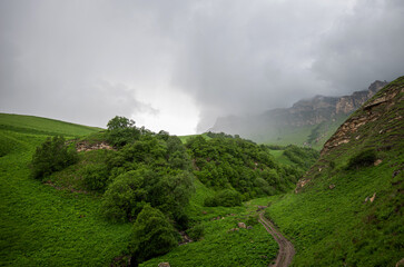 Beautiful spring landscape in the Caucasus Mountains with lush grassy hills and rocky slopes. Dramatic cloudy sky on a rainy day. Low clouds on the tops of the mountains.