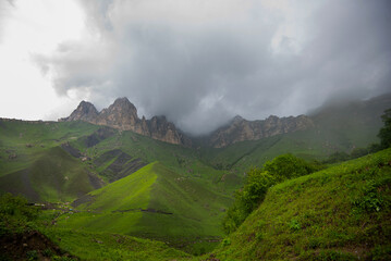 Beautiful spring landscape in the Caucasus Mountains with lush grassy hills and rocky slopes. Dramatic cloudy sky on a rainy day. Low clouds on the tops of the mountains.