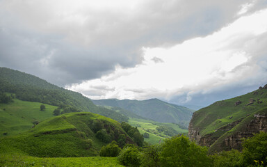 Fototapeta premium Beautiful spring landscape in the Caucasus Mountains with lush grassy hills and rocky slopes. Dramatic cloudy sky on a rainy day. Low clouds on the tops of the mountains.