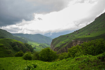 Naklejka premium Beautiful spring landscape in the Caucasus Mountains with lush grassy hills and rocky slopes. Dramatic cloudy sky on a rainy day. Low clouds on the tops of the mountains.