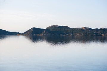 A lake with morning haze, calm water and reflection. Idyllic lake view. There are mountain ranges around the lake. The calm surface of the lake. There is a dense forest on the shore.