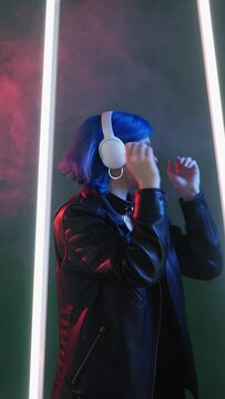 Playlist joy. Techno music. Cyber gadget. Satisfied dancing woman listening song in futuristic earphones in pink neon LED light on smoke dark background vertical.