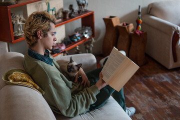 Blond boy enjoys reading a book on the sofa in the living room of his house and his cat accompanies him in a relaxed way