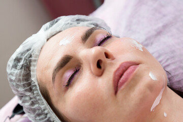 Obraz na płótnie Canvas An adult brunette woman with her eyes closed after facial rejuvenation procedure with cream on her face.