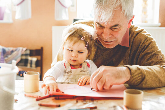 Cute little baby toddler girl and handsome senior grandfather painting with colorful pencils at home. Grandchild and man having fun together. Family and generation in love