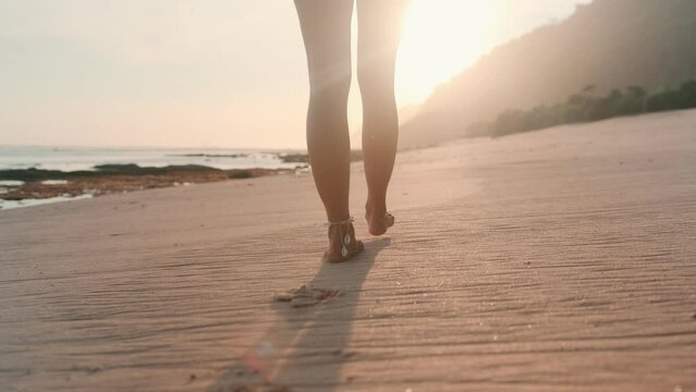 Young beautiful Caucasian woman in swimsuit slowly leaving footprints in sand walks with back to camera enjoying sun's rays goes towards sunset wander along beach. Travel, summer tourism concept