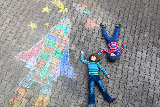 Two little kids boys having fun with drawing universe and space shuttle picture with colorful chalks. Creative leisure for children outdoors in summer. Siblings, friends playing and painting on ground
