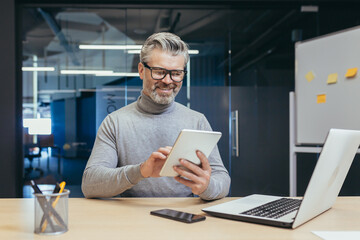 Successful gray-haired businessman working in modern office, man holding tablet computer in hands rejoices and smiles reads news