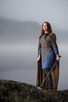 Red-haired girl in armor with sword and raincoat on river bank