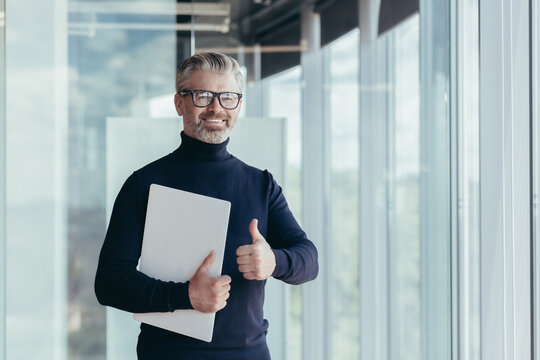 Portrait of senior and experienced teacher, gray-haired university teacher by the window smiling and looking at camera holding laptop in hand and showing thumbs up approvingly