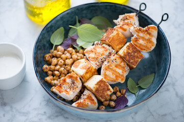 Grilled chicken meat and halloumi cheese on skewers served in a blue bowl, middle close-up on a light-grey marble background