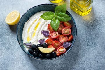 Bowl of greek tarama dip with black olives, cherry tomatoes and fresh basil, elevated view on a light-blue stone background, studio shot