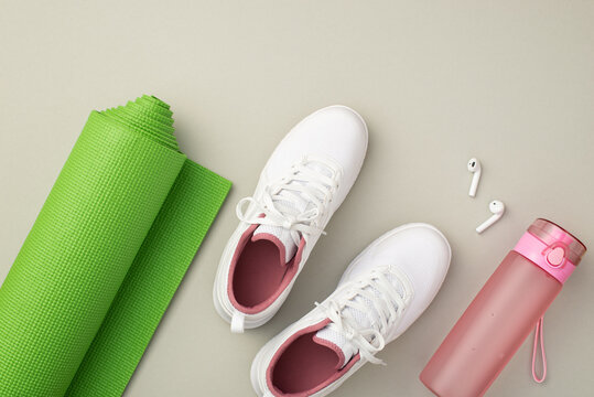 Fitness concept. Top view photo of white sports shoes pink bottle of water earbuds and green exercise mat on isolated pastel grey background