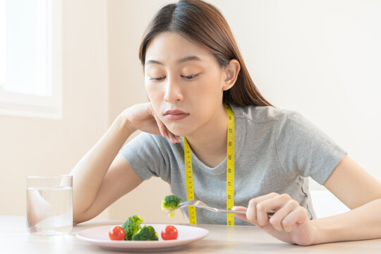 Anorexia, unhappy beautiful asian young woman, girl on dieting, holding fork at broccoli in salad plate, dislike or tired with eat fresh vegetables. Nutrition of good healthy, bored with food.