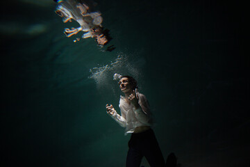 Fototapeta na wymiar underwater shooting with contrasting light, a guy in a white shirt and pants screaming underwater, panicking and afraid of drowning, falling into the water, a crime.