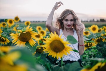 Fototapeta na wymiar A young, slender girl with loose hair in a T-shirt stands in a field of sunflowers at sunset