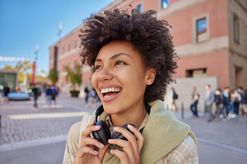 Fototapeta Outdoor shot of cheerful woman with curly hair dressed in casual clothes wears stereo headphones around neck has glad expression walks at busy street against blurred background. Lifestyle concept obraz
