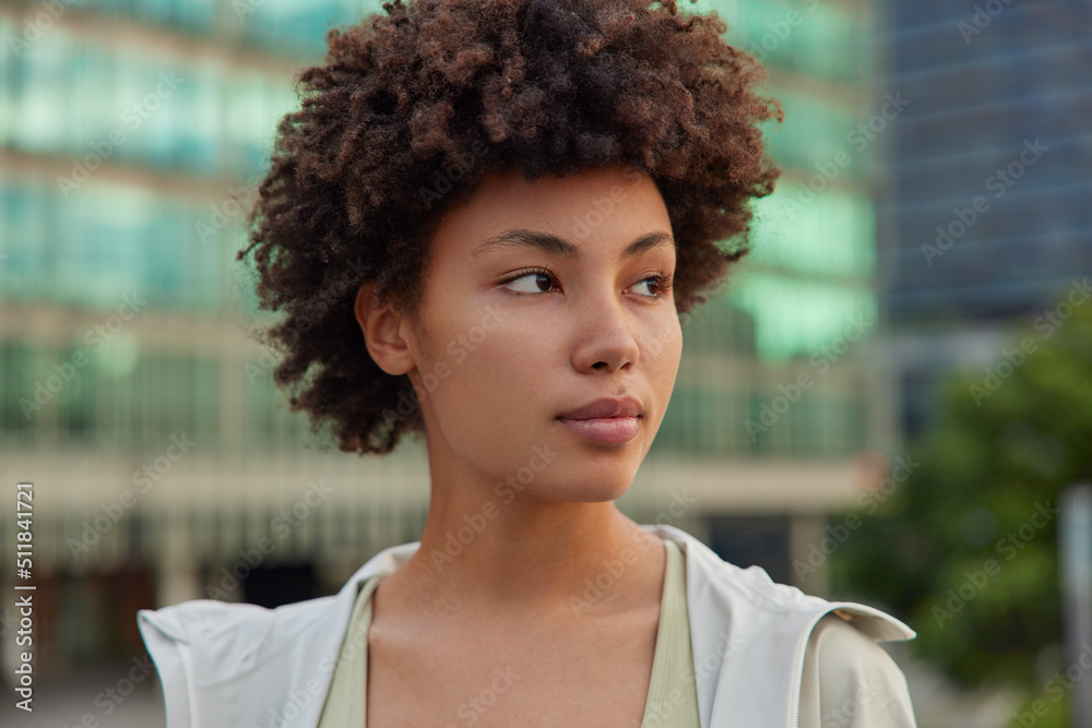 Wall mural outdoor shot of pensive curly haired young woman concentrated away strolls at street dressed in stre