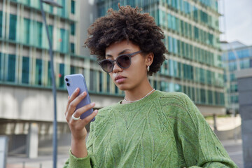 Fototapeta na wymiar Photo of stylish curly haired young woman wears sunglasses and green jumper checks received email message browses website in social media stands outdoors against modern buildings on urban street