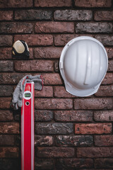 A red construction level, measuring tape measure, gloves and a white helmet on a brick wall.