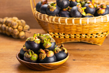 Pile of fresh mangosteen fruit in wooden plate and in bamboo basket with bunch of longkong on wooden table.