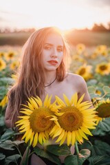 A young, naked, slender girl with loose hair covers her body with sunflowers in a field at sunset