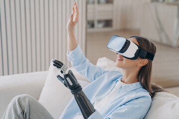 Happy disabled european girl with bionic cyber arm in virtual reality glasses at home.