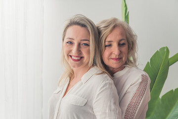 Lovely caucasian blue-eyed blonde millenial women standing near a window in a bright apartment hugging each other. High quality photo