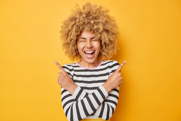 Happy young woman with curly blonde hair laughs out gladfully points sideways selects between two options picks two variants dressed in casual striped jumper isolated over yellow background.