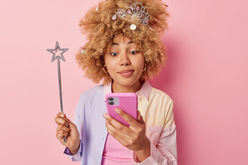Lovely curly haired woman checks received message on smartphone uses modern gadget holds magic wand dresses for costume party wears crown and shirt isolated over pink background. Festive occasion © WHstudio Leushin N
