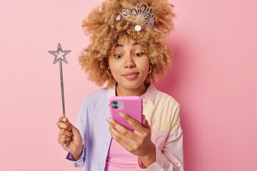 Lovely curly haired woman checks received message on smartphone uses modern gadget holds magic wand...