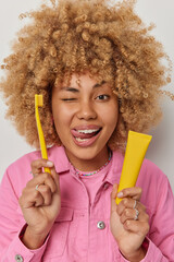 Vertical shot of cheerful curly haired young woman sticks out tongue winks eye recommends good toothpaste for your teeth dressed in pink jacket isolated over white background. Tooth care concept