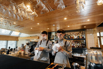 Two stylish bartenders in masks and uniforms during the pandemic, rub glasses to shine. The work of restaurants and cafes during the pandemic..