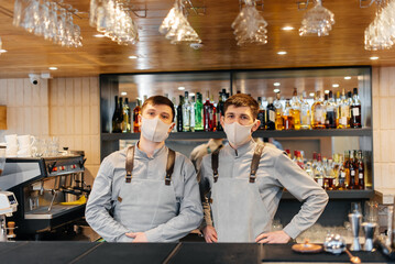 Two stylish bartenders in masks and uniforms during the pandemic, stand behind the bar. The work of restaurants and cafes during the pandemic.