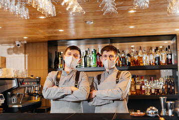 Two stylish bartenders in masks and uniforms during the pandemic, stand behind the bar. The work of restaurants and cafes during the pandemic.