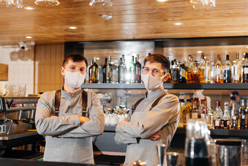 Two stylish bartenders in masks and uniforms during the pandemic, stand behind the bar. The work of restaurants and cafes during the pandemic.