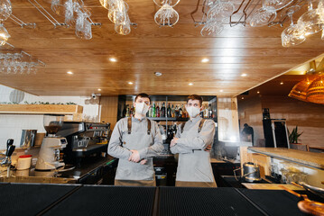 Two stylish bartenders in masks and uniforms during the pandemic, stand behind the bar. The work of restaurants and cafes during the pandemic.