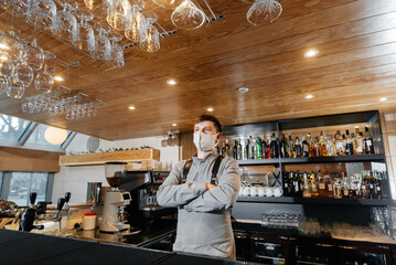 Fototapeta na wymiar A stylish bartender in a mask and uniform stands near the bar during the pandemic. The work of restaurants and cafes during the pandemic.