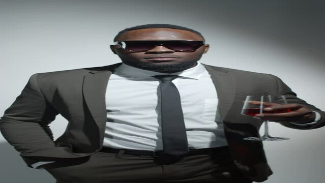 Vertical format footage of young African American man in elegant suit and trendy sunglasses holding glass of wine and posing for camera with smile in studio