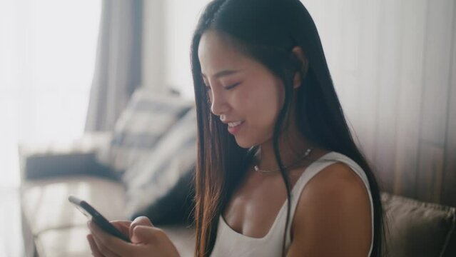 Happy young asian woman holding smart phone laughing enjoying using mobile sitting at home at sunlight. Portrait. Slow motion