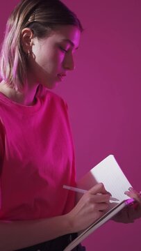 To do list. Personal development. Goal motivation. Mindful dreamy woman writing task note idea in journal in vivid neon light isolated on pink background vertical.