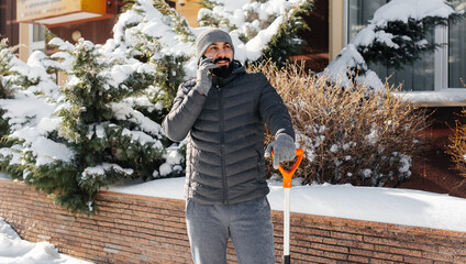 A man stands with a shovel and removes snow in front of the house on a sunny and frosty day and talks on the phone. Cleaning the street from snow on a winter day.  © Andrii