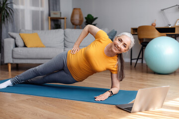 Happy fit senior woman standing in side plank, training on yoga mat in living room and using laptop