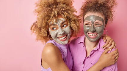 Horizontal shot of cheerful friendly young women hug and look happily away have curly hair apply beauty masks for skin treatment isolated over pink background blank space for your advertisement
