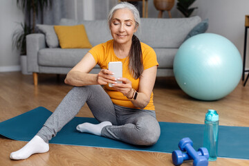 Happy athletic senior woman in sportswear sitting on yoga mat at home and using modern cellphone